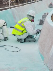 Worker in a bright safety vest and hard hat kneels by a curved pool wall, using a tool to smooth turquoise surface.