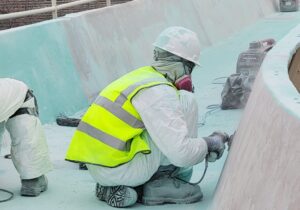 Worker in a bright safety vest and hard hat kneels by a curved pool wall, using a tool to smooth turquoise surface.